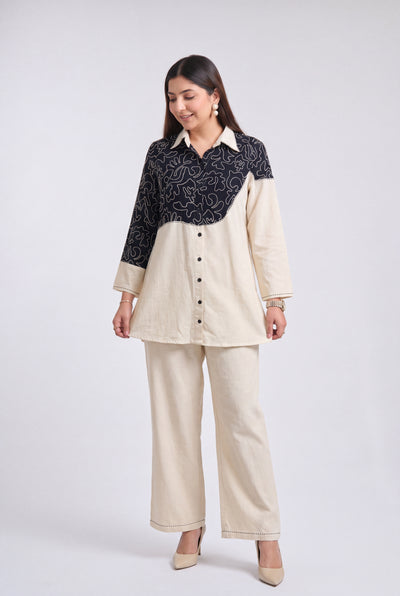A woman modeling a two-piece outfit consisting of a color-blocked button-up shirt with a black and beige floral pattern on the upper half and solid beige on the lower half, paired with matching beige wide-leg trousers, against a light gray studio background.