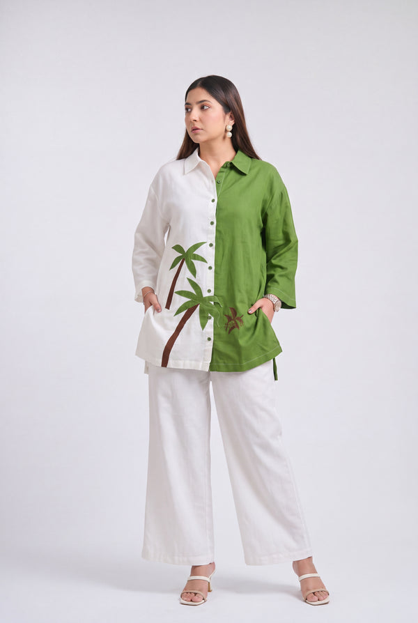 A woman modeling a two-tone button-up shirt with white on one side and olive green on the other, featuring a palm tree embroidery on the white side, paired with white wide-leg pants against a light gray studio background.