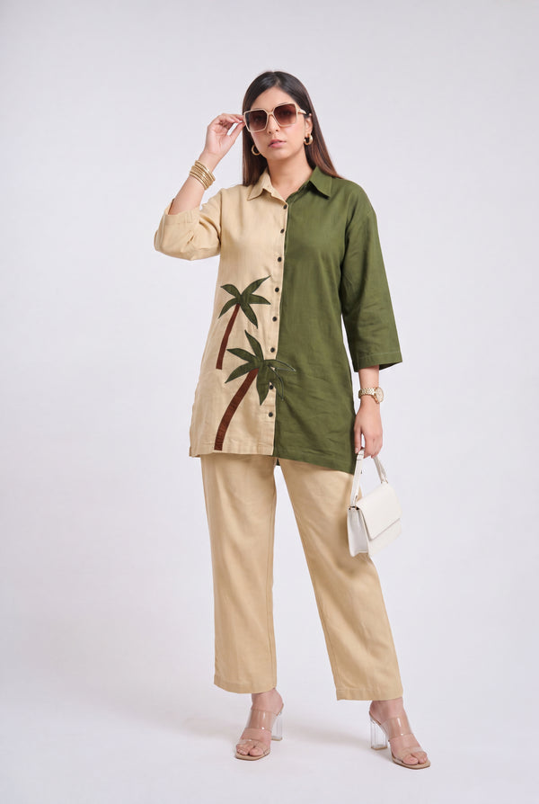 A woman models a two-tone button-up shirt and matching wide-leg trousers. The shirt is split vertically with one side in beige featuring a green palm tree print and the other side in olive green.