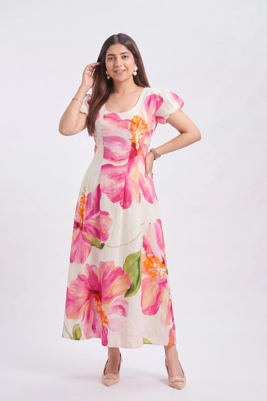 A woman modeling a short-sleeve, scoop-neck maxi dress with a vibrant pink and orange floral print on a cream background, set against a plain light gray studio backdrop.
