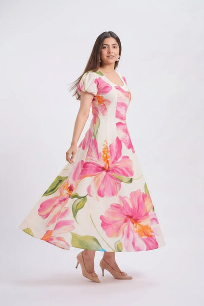 A woman modeling a short-sleeve V-neck maxi dress with a vibrant pink and orange hibiscus floral print on a cream background, set against a plain light gray studio backdrop.