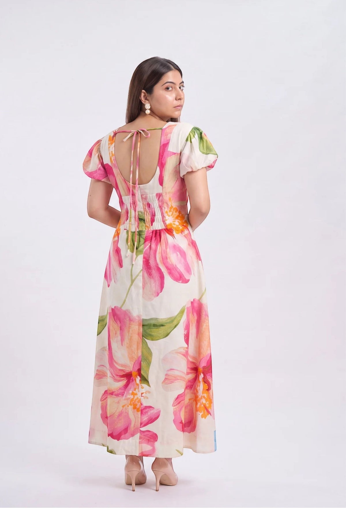 A woman modeling a long-sleeved maxi dress from the back, featuring a vibrant pink and green floral print on a cream background with a tie-back neckline, set against a plain light gray studio backdrop.