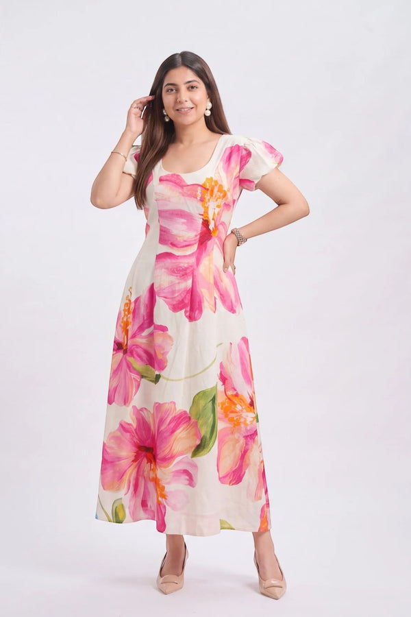 A woman modeling a short-sleeve, scoop-neck maxi dress with a vibrant pink and orange floral print on a cream background, set against a plain light gray studio backdrop.