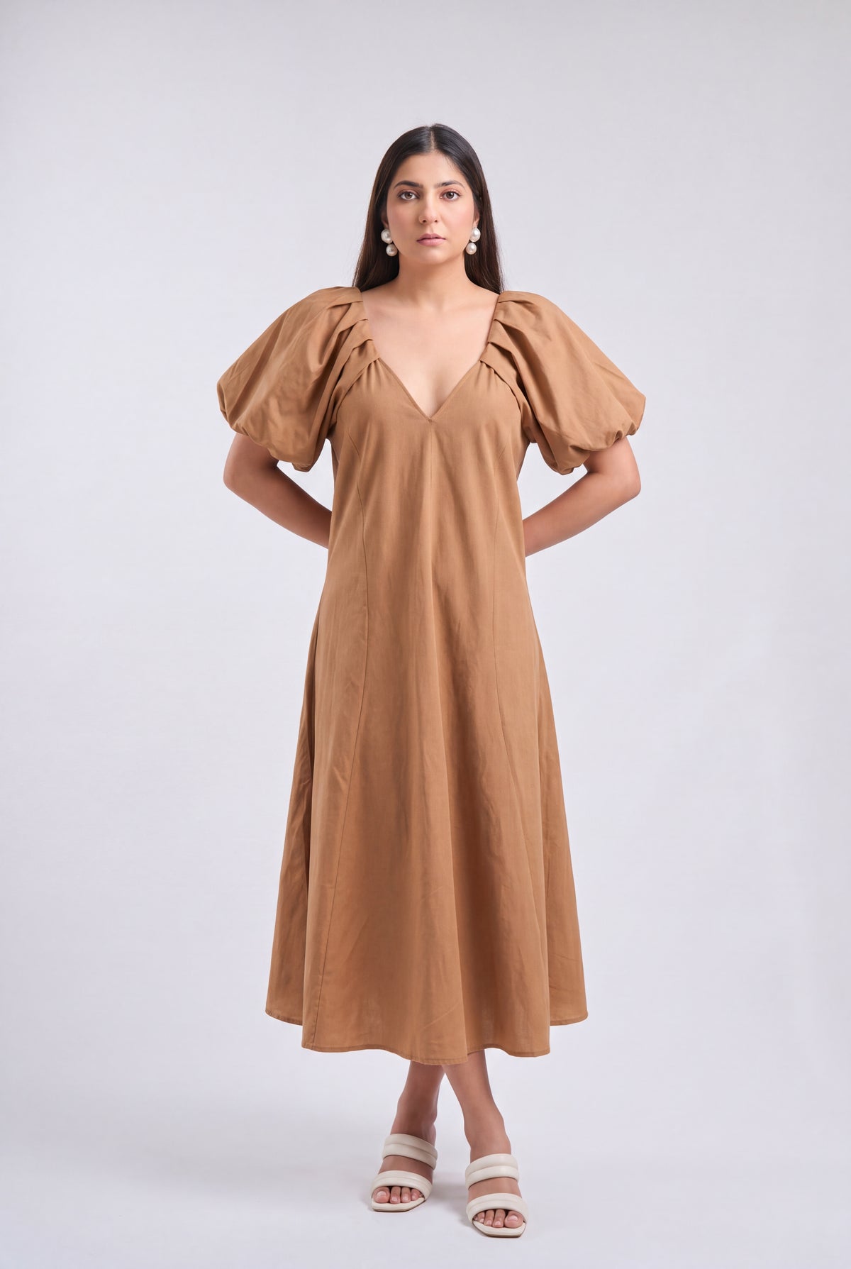 A woman modeling a tan midi dress with voluminous puff sleeves, a deep V-neckline, and a flowing skirt, paired with white heeled sandals against a light gray studio background.