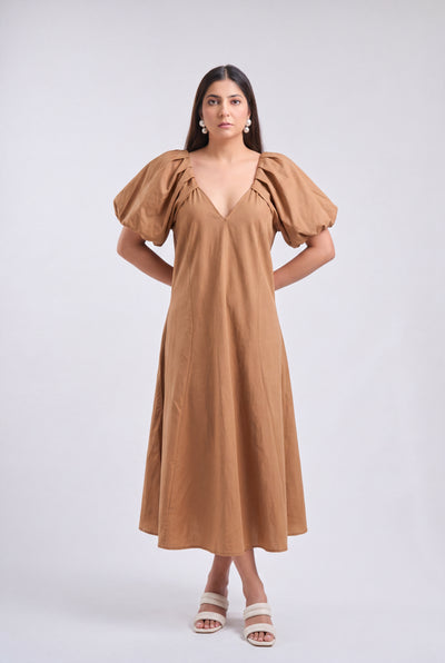 A woman modeling a tan midi dress with voluminous puff sleeves, a deep V-neckline, and a flowing skirt, paired with white heeled sandals against a light gray studio background.
