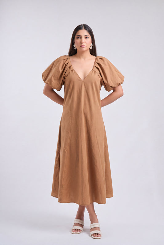 A woman modeling a tan midi dress with voluminous puff sleeves, a deep V-neckline, and a flowing skirt, paired with white heeled sandals against a light gray studio background.