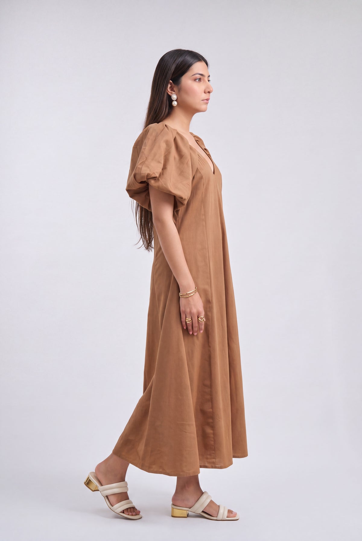 A woman modeling a brown midi dress with puff sleeves and a V-neckline, standing in profile against a light gray studio background.
