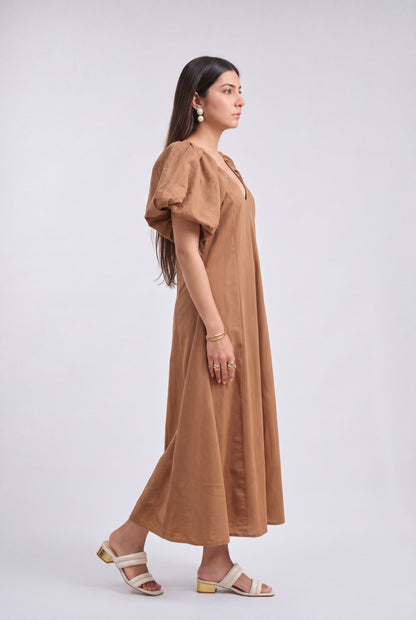 A woman modeling a brown midi dress with puff sleeves and a V-neckline, standing in profile against a light gray studio background.