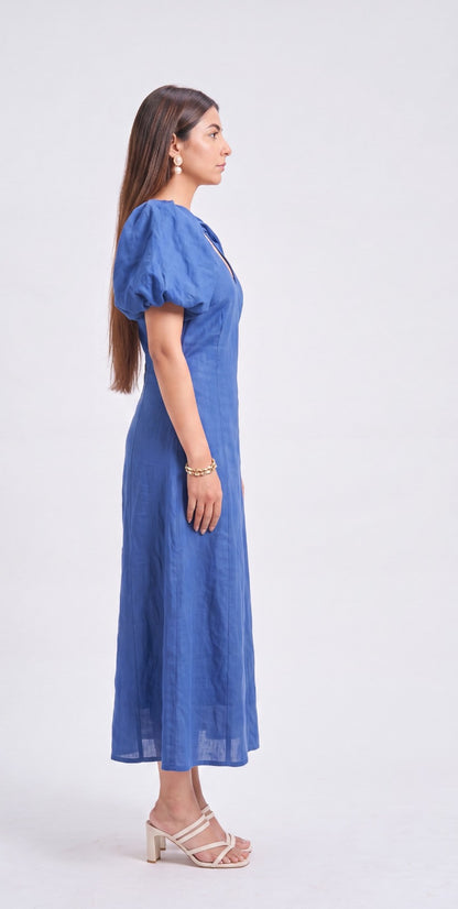 A woman modeling a blue midi dress with puffed short sleeves, a V-neckline, and a flowing skirt, shown in profile against a plain light gray studio background.