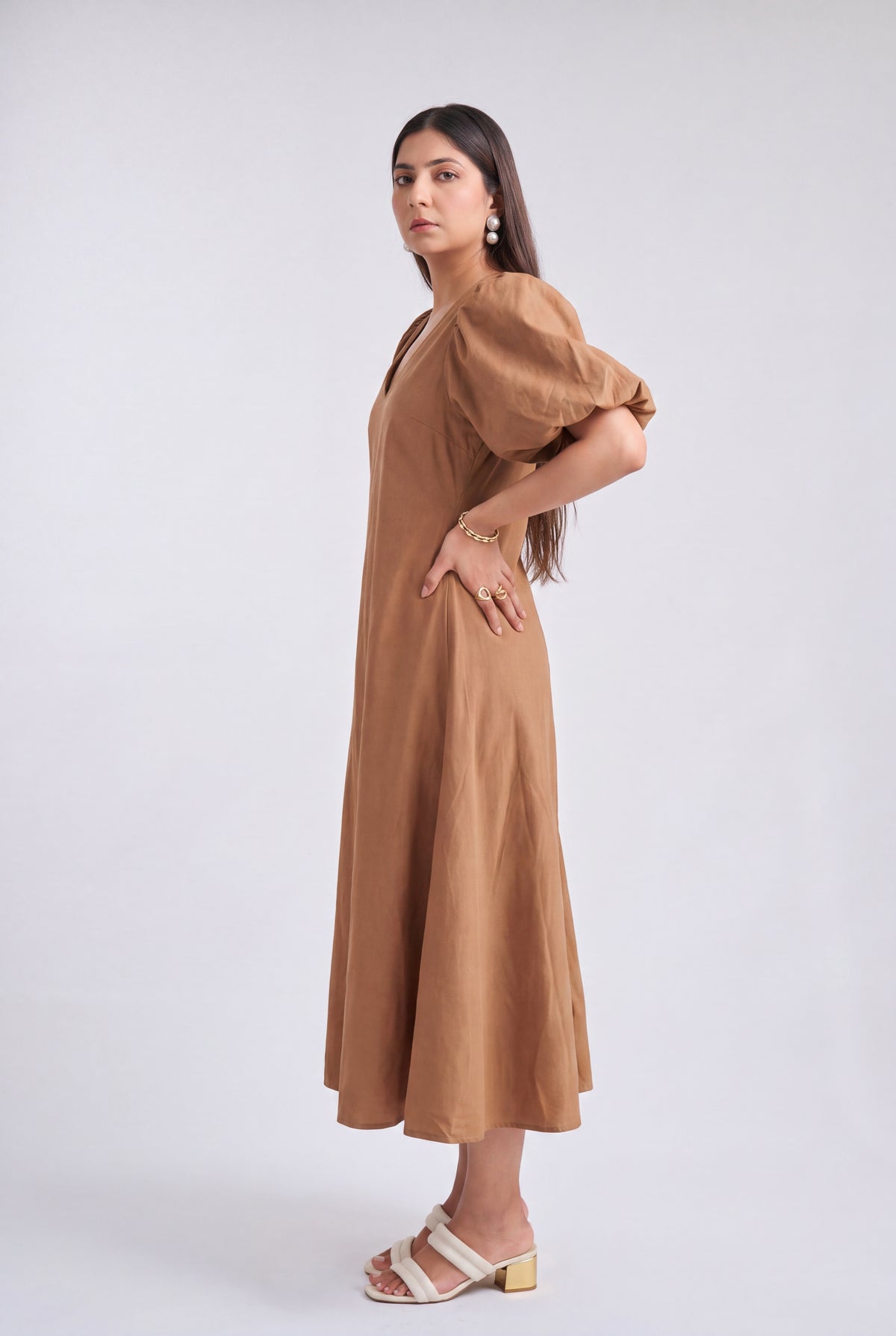 A woman modeling a brown midi dress with puff sleeves and a V-neckline, paired with white heeled sandals, against a light gray studio background.
