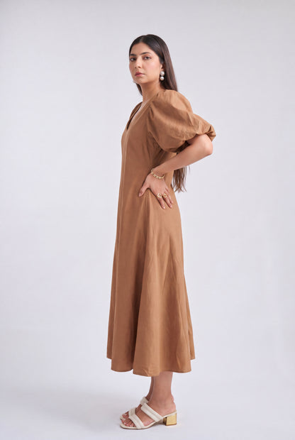 A woman modeling a brown midi dress with puff sleeves and a V-neckline, paired with white heeled sandals, against a light gray studio background.