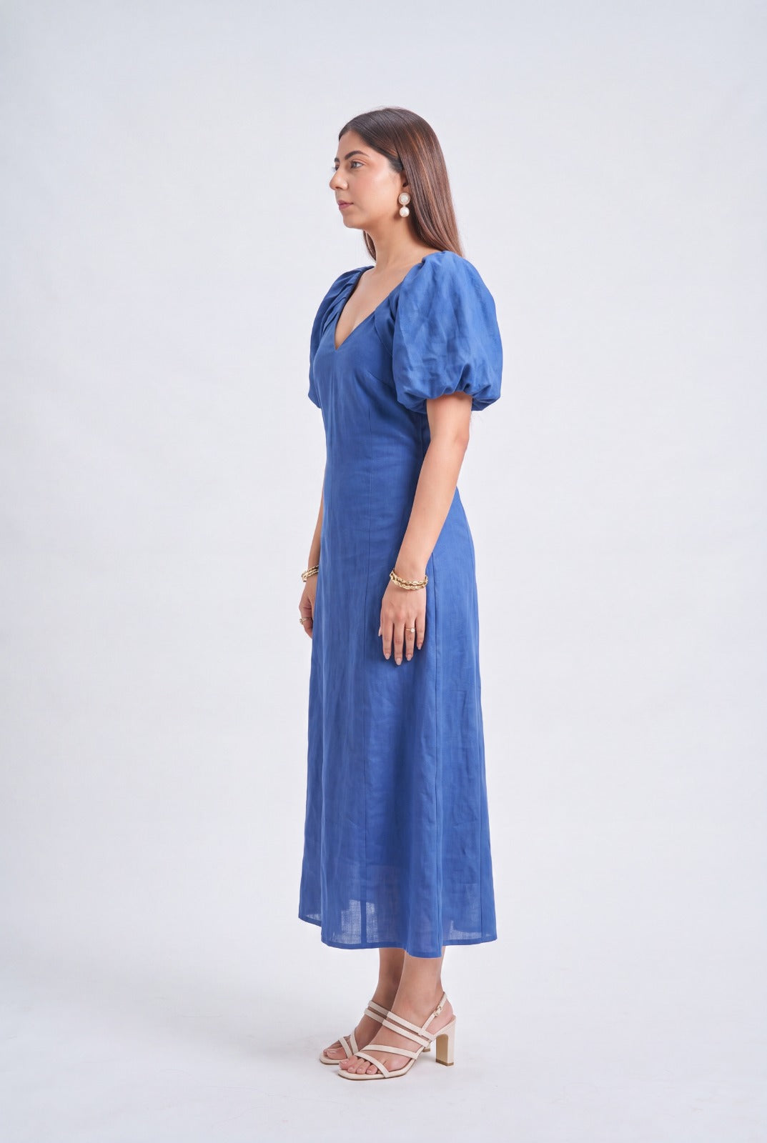A woman modeling a blue midi dress with puff sleeves and a V-neckline, standing in profile against a light gray studio background.