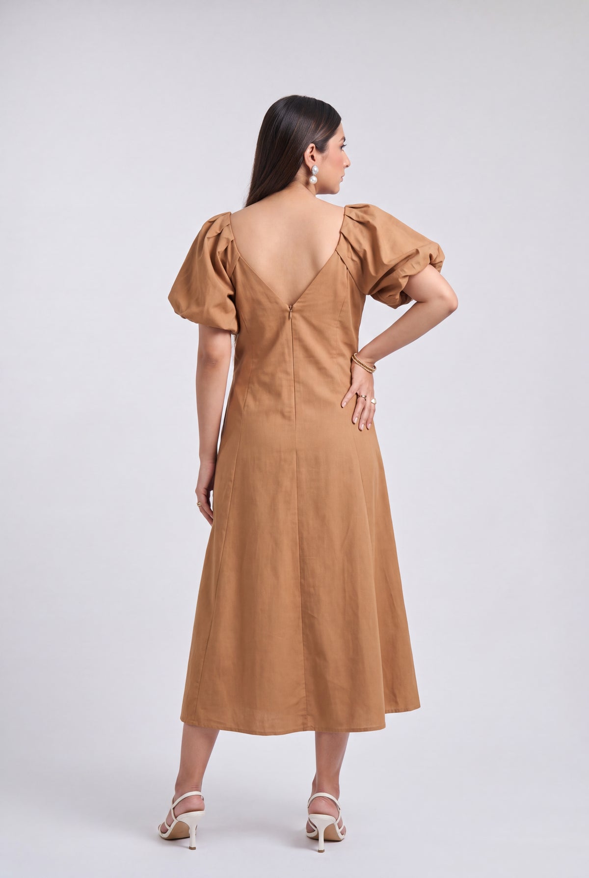 A woman is shown from the back wearing a tan midi dress with puff sleeves and a deep V-back, paired with beige high-heeled sandals, against a light gray studio background.