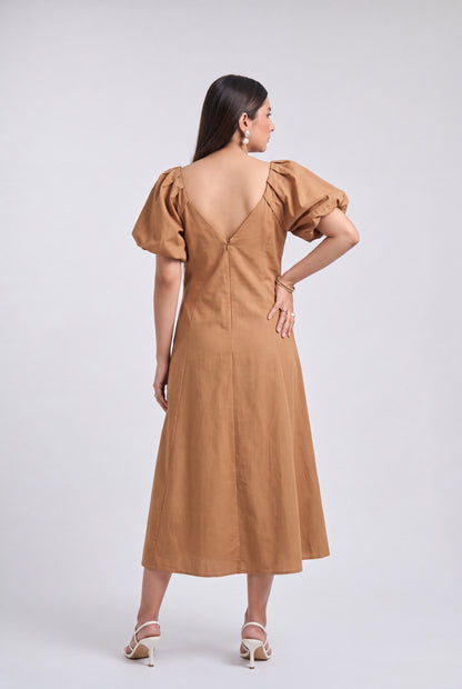 A woman is shown from the back wearing a tan midi dress with puff sleeves and a deep V-back, paired with beige high-heeled sandals, against a light gray studio background.