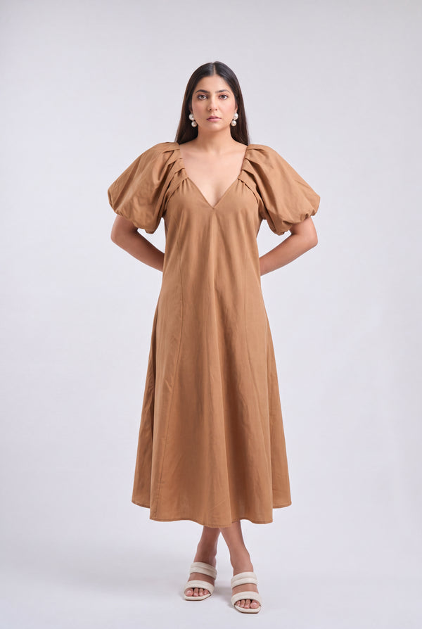 A woman modeling a tan midi dress with voluminous puff sleeves, a deep V-neckline, and a flowing skirt, paired with white heeled sandals against a light gray studio background.