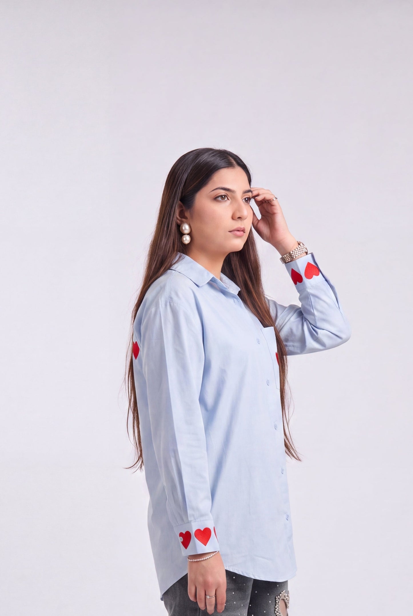 A woman modeling a light blue long-sleeve button-up shirt with red heart embroidery on the cuffs and shoulders, paired with distressed gray jeans and beige heeled sandals, against a plain light gray studio background.