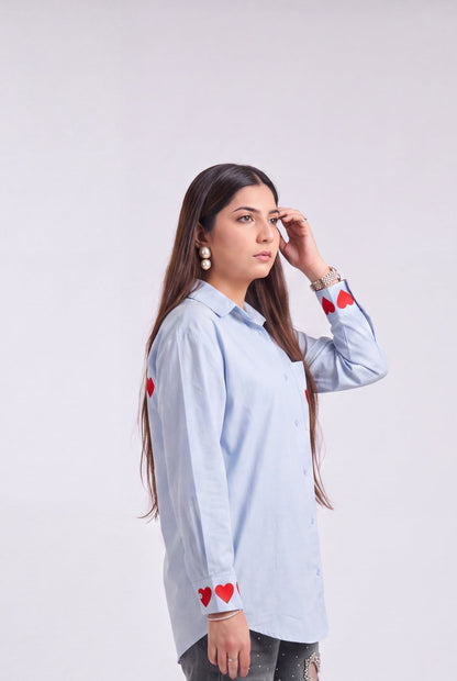 A woman modeling a light blue long-sleeve button-up shirt with red heart embroidery on the cuffs and shoulders, paired with distressed gray jeans and beige heeled sandals, against a plain light gray studio background.