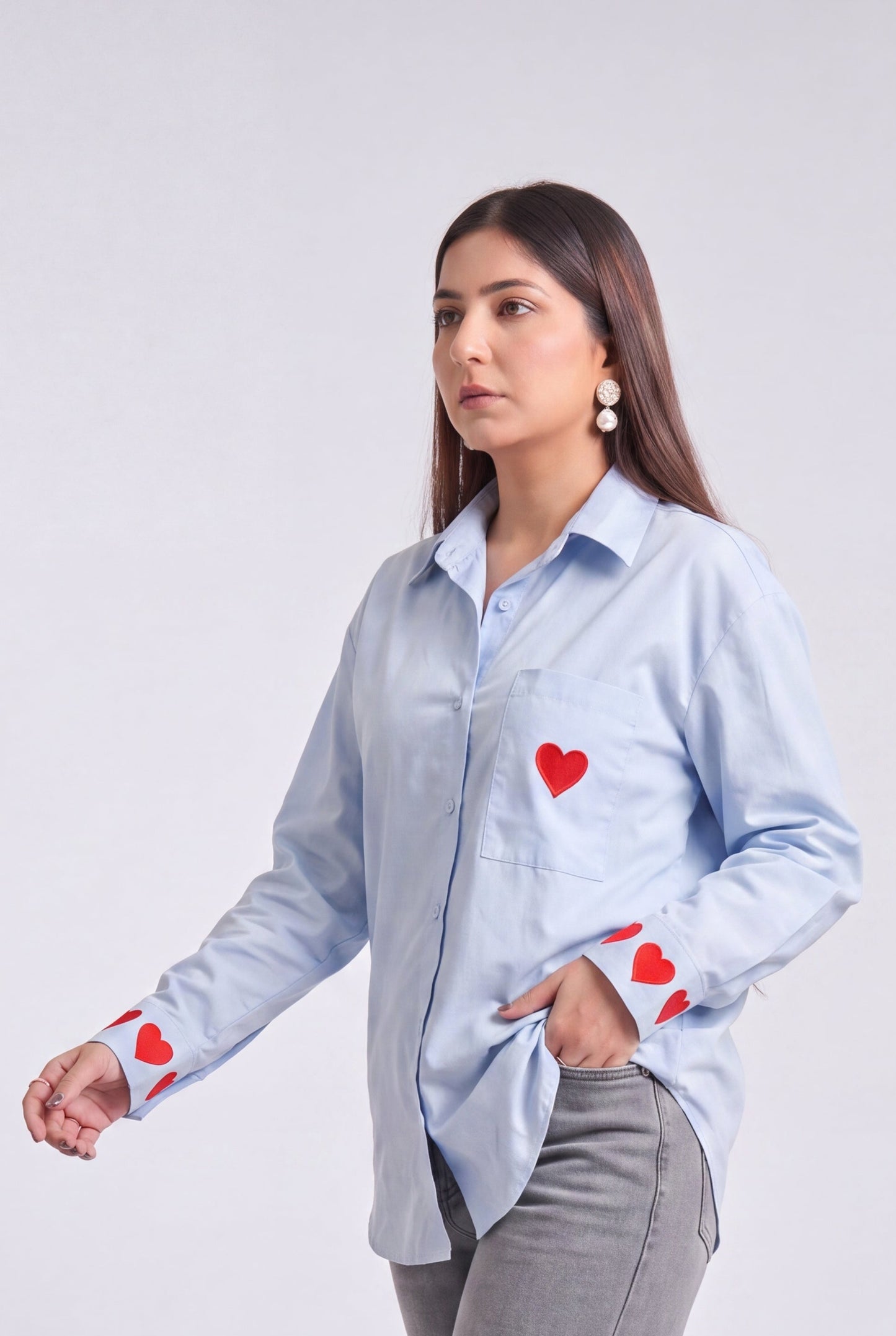 A woman modeling a light blue button-up shirt with red heart details on the chest and cuffs, paired with gray wide-leg jeans and white heeled sandals, against a light gray studio background.