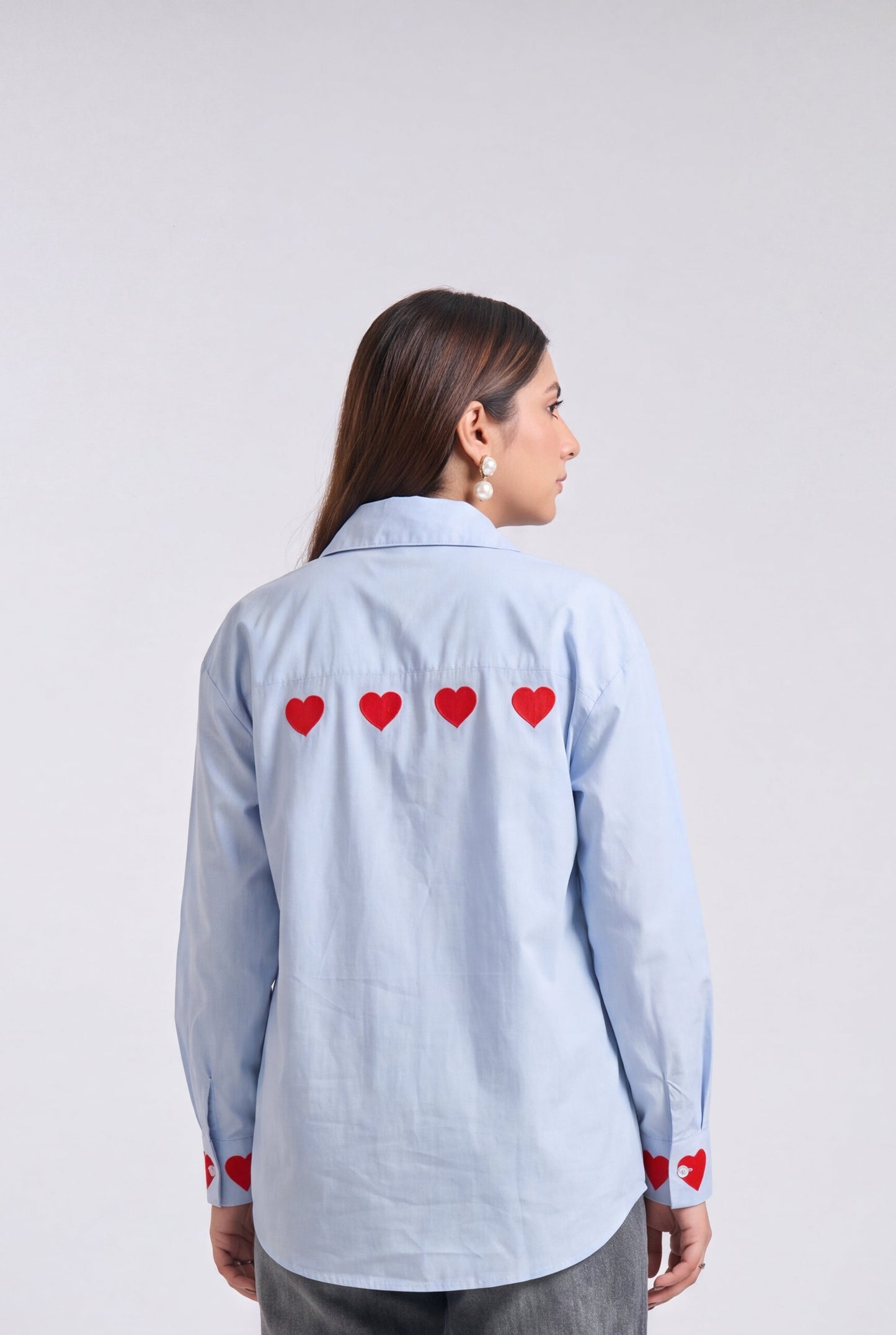 A woman shown from the back wearing a light blue long-sleeve button-up shirt with four red embroidered hearts across the upper back and distressed gray jeans with heart patches on the knees, set against a plain light gray studio background.
