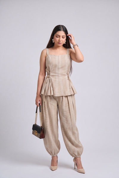 A woman modeling a beige sleeveless peplum top and matching wide-leg pants set, holding a small black handbag, against a light gray studio background.
