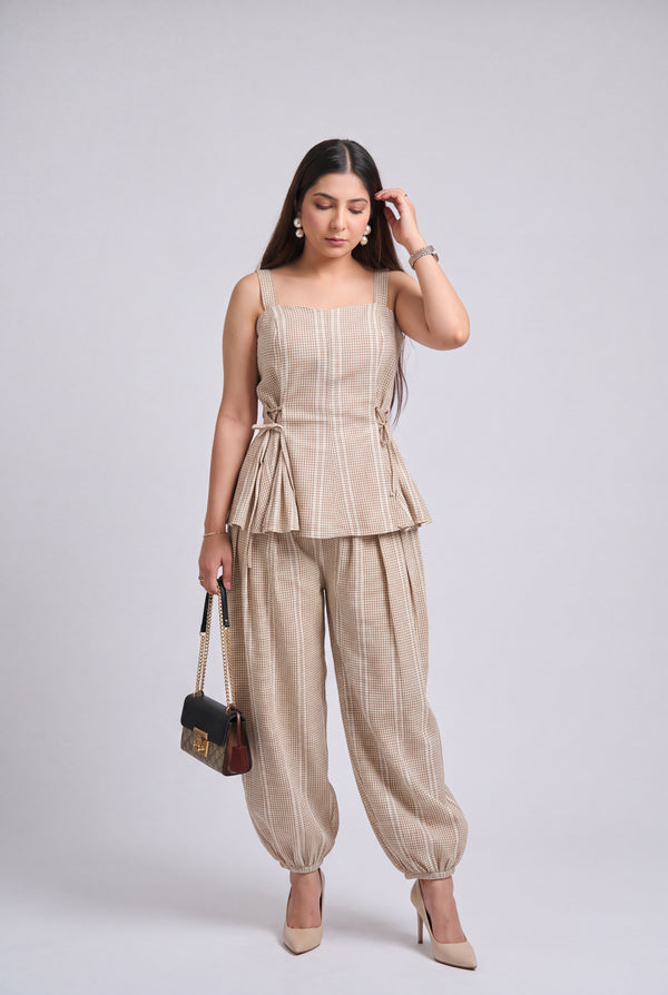 A woman modeling a beige sleeveless peplum top and matching wide-leg pants set, holding a small black handbag, against a light gray studio background.