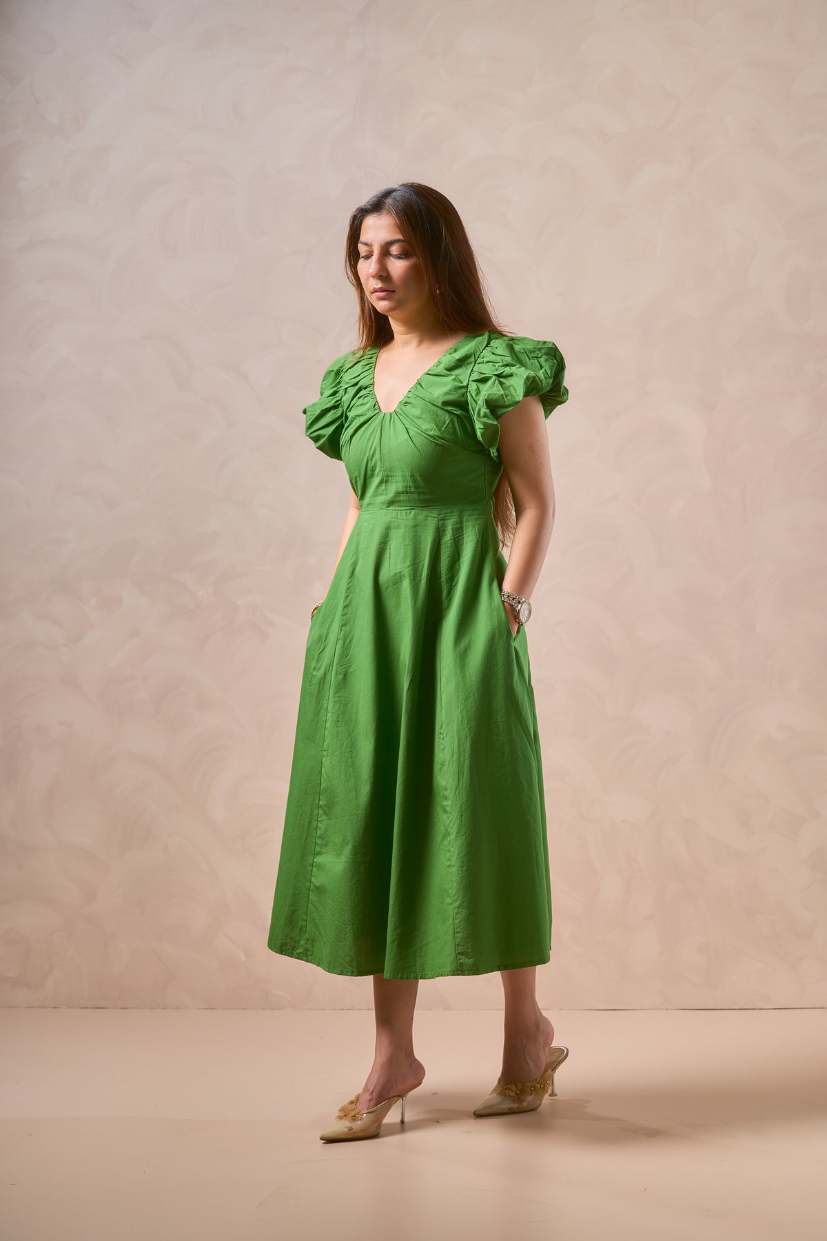 A woman modeling a bright green midi dress with ruffled short sleeves, a gathered waist, and side pockets, standing against a textured beige studio backdrop.
