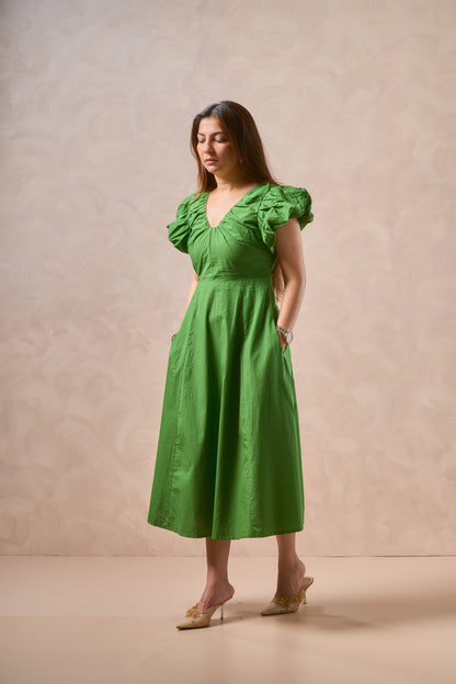 A woman modeling a bright green midi dress with ruffled short sleeves, a gathered waist, and side pockets, standing against a textured beige studio backdrop.
