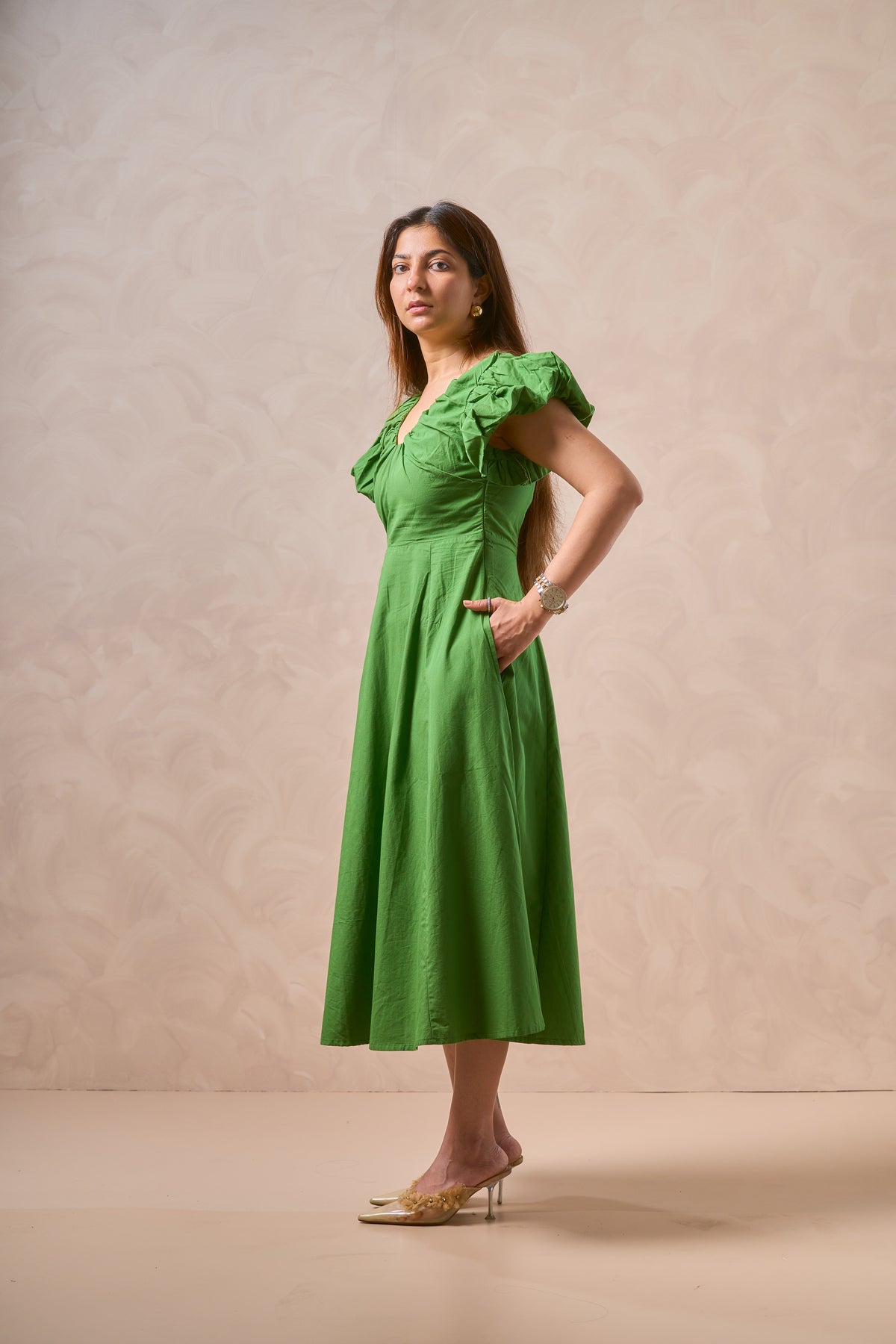 A woman modeling a bright green midi dress with ruffled short sleeves, a gathered waist, and a flared skirt, standing against a textured beige studio backdrop.