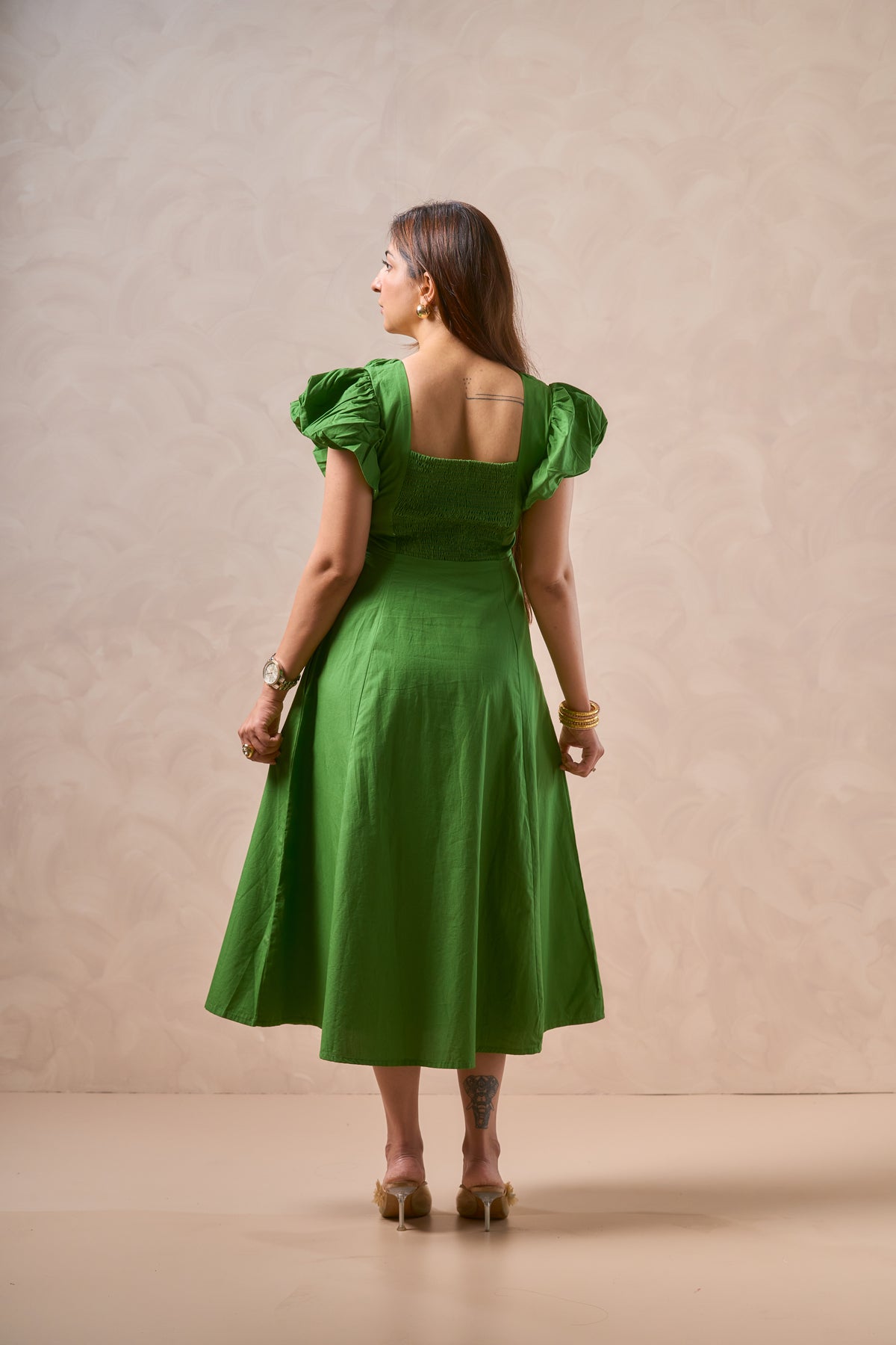 A woman stands with her back to the camera wearing a bright green midi dress with puffed short sleeves, a square neckline, and a flared skirt. She is posed against a textured beige studio backdrop.