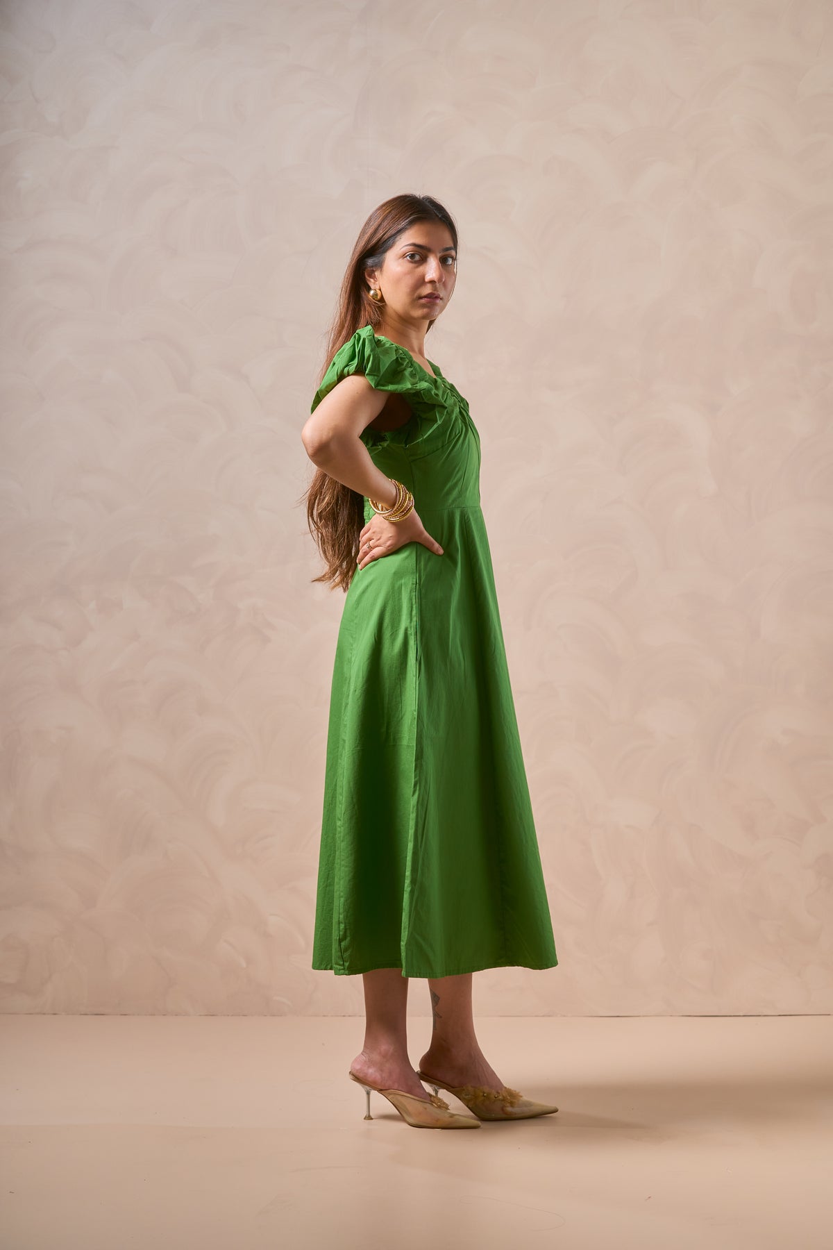 A woman modeling a green midi dress with ruffled sleeves, standing against a textured beige studio backdrop.