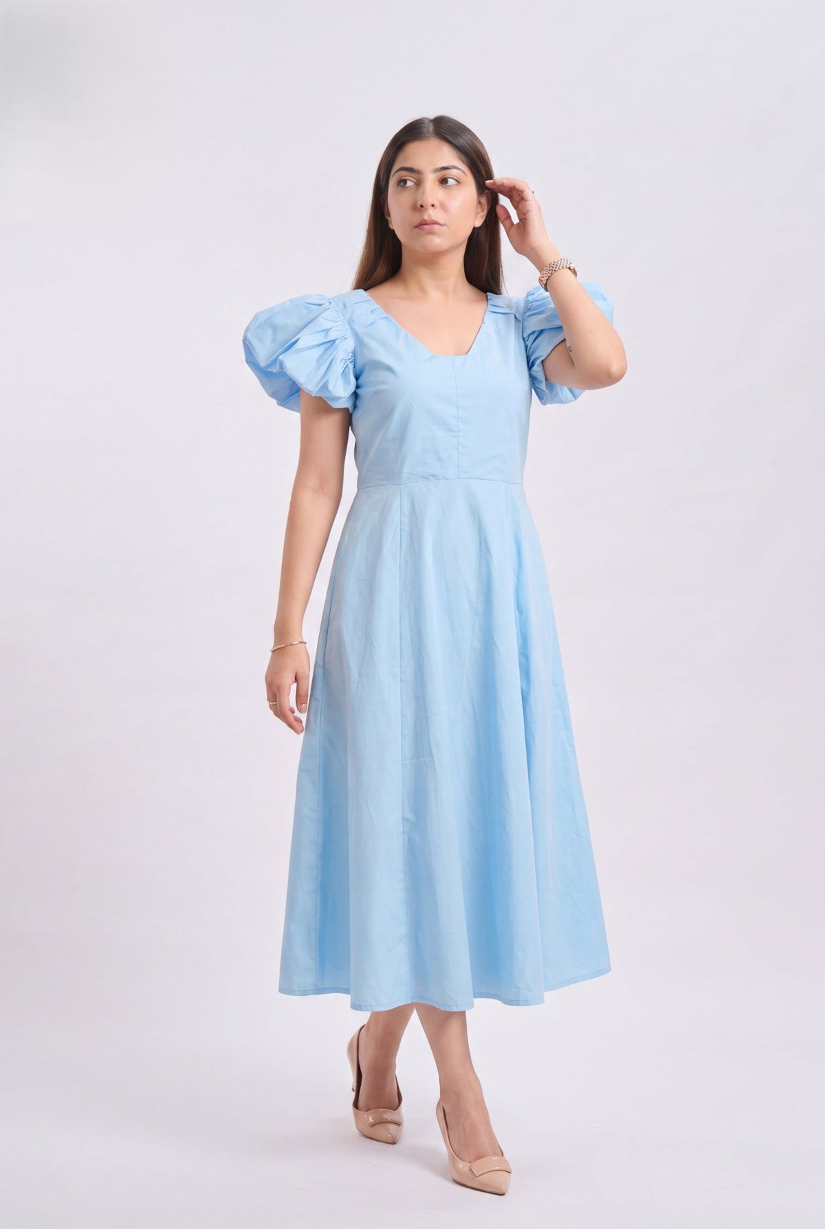 A woman modeling a light blue midi dress with puffed short sleeves, a V-neckline, and a flared skirt, photographed against a plain light gray studio background.