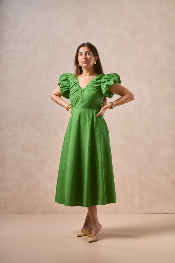 A woman modeling a vibrant green midi dress with ruffled puff sleeves, a gathered sweetheart neckline, and a flared skirt, standing against a textured beige studio backdrop.