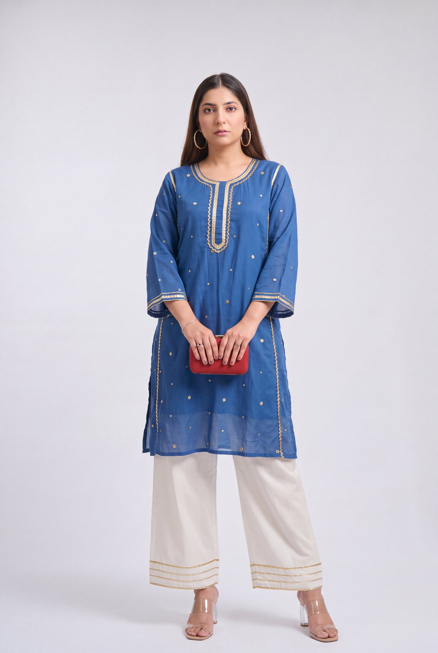 A woman modeling a blue kurta with gold embroidery, paired with off-white wide-leg pants and holding a small red clutch, against a light gray studio background.