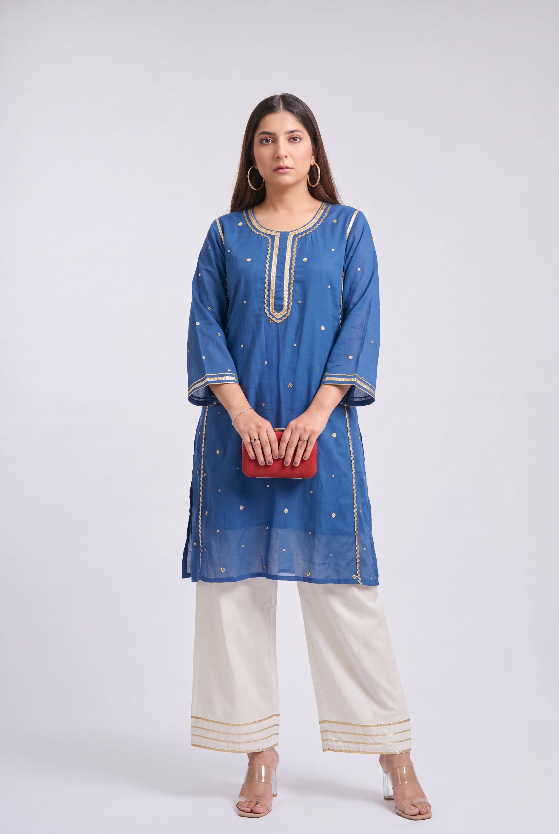 A woman modeling a blue kurta with gold embroidery, paired with off-white wide-leg pants and holding a small red clutch, against a light gray studio background.