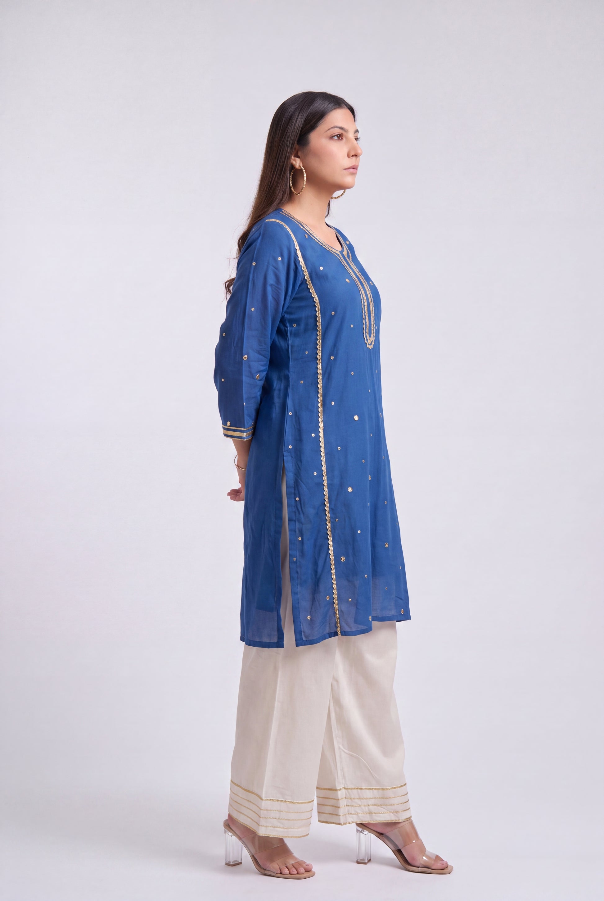 A woman modeling a blue kurta with gold embroidery, paired with cream wide-leg pants and clear high-heeled sandals, photographed against a light gray studio background.