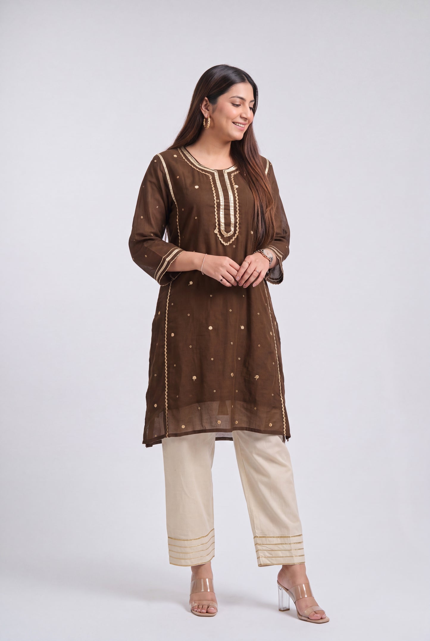 A woman modeling a brown embroidered kurta with beige pants and clear high-heeled sandals, standing against a light gray studio background.