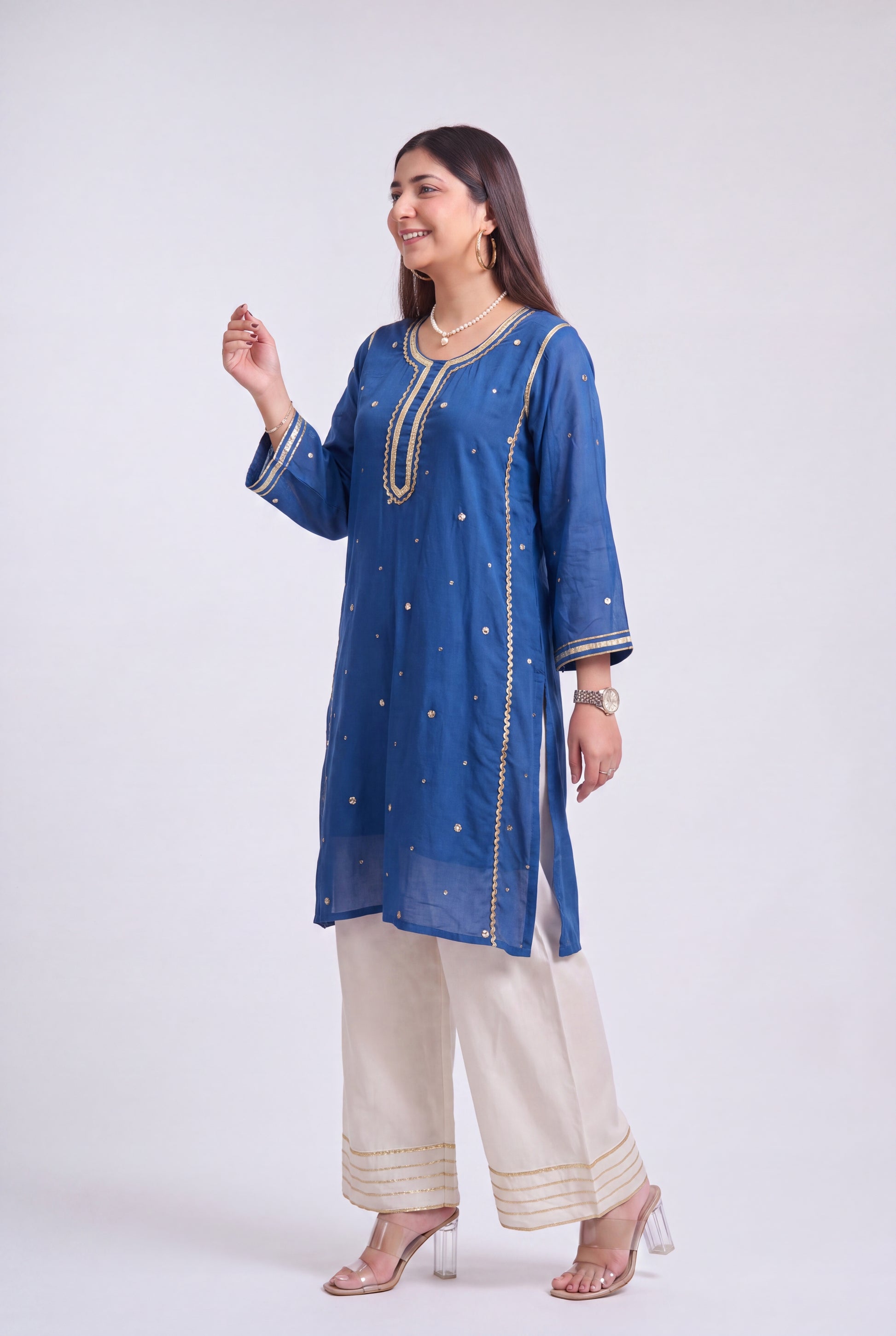 A woman modeling a blue embroidered kurta with beige wide-leg pants and clear high-heeled sandals, standing against a light gray studio background.