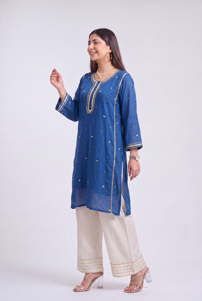 A woman modeling a blue embroidered kurta with beige wide-leg pants and clear high-heeled sandals, standing against a light gray studio background.