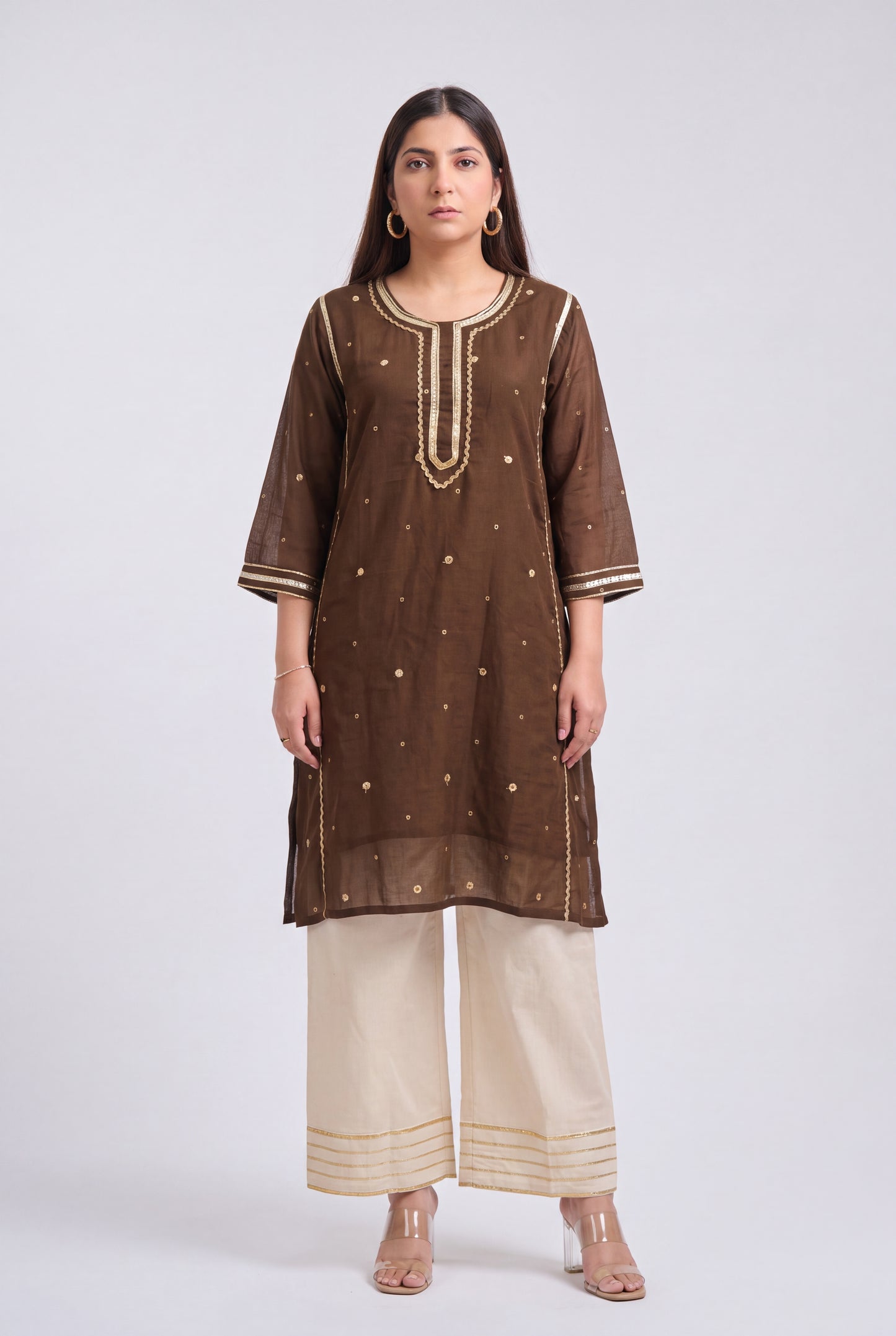 A woman modeling a brown sheer embroidered kurta with gold detailing, paired with cream wide-leg pants, against a light gray studio background.