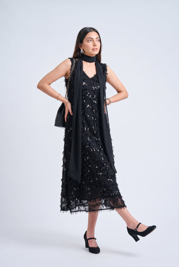 A woman modeling a black sleeveless lace midi dress with sequined details, a matching black choker, and a draped black scarf, paired with black Mary Jane heels against a light gray studio background.