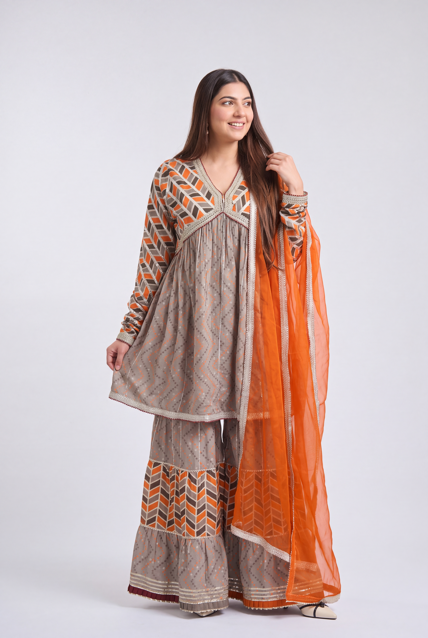 A woman modeling a traditional South Asian outfit consisting of a beige kurta and wide-leg pants with orange and brown geometric patterns, paired with a bright orange dupatta, against a light gray studio background.