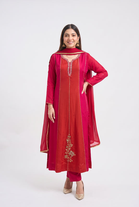 A woman modeling a traditional Indian salwar kameez set in a vibrant red and magenta colorway, featuring gold embroidery on the front and a matching sheer dupatta, set against a plain white studio background.