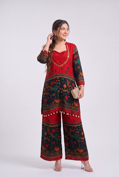 A woman modeling a red and black printed kurta set with wide-leg pants, gold embroidery, and a small beige clutch, photographed against a light gray studio background.