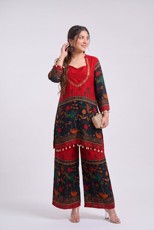 A woman modeling a red and black printed kurta set with wide-leg pants, gold embroidery, and a small beige clutch, photographed against a light gray studio background.