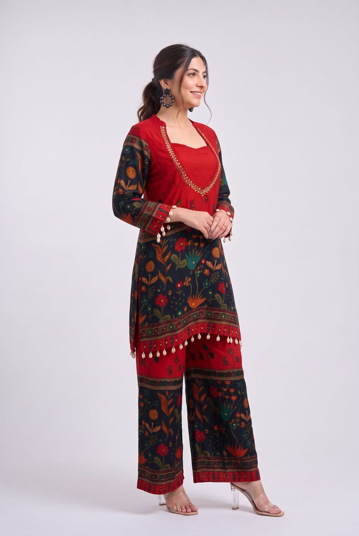 A woman modeling a traditional Indian salwar suit set featuring a red kurta with gold embroidery and a matching printed trouser set in red and black with floral motifs and tassel trim, set against a plain light gray background.
