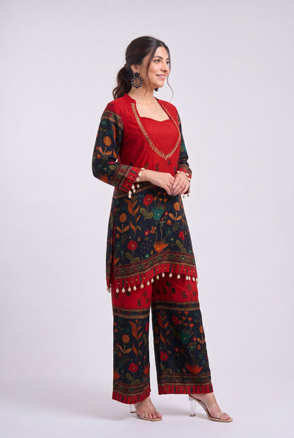 A woman modeling a traditional Indian salwar suit set featuring a red kurta with gold embroidery and a matching printed trouser set in red and black with floral motifs and tassel trim, set against a plain light gray background.