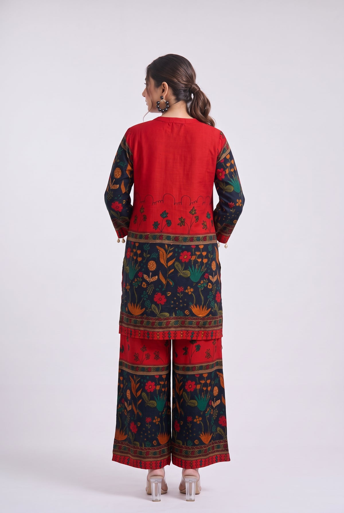 A woman is shown from the back wearing a traditional Indian outfit consisting of a red kurta with floral patterned sleeves and a matching pair of wide-leg trousers. The outfit features a vibrant red base with dark navy blue sleeves and pant legs decorated with colorful floral motifs in orange, green, and yellow.