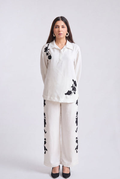 A woman modeling a matching two-piece outfit consisting of a long-sleeve collared top and wide-leg trousers in off-white, both featuring black floral embroidery, set against a plain light gray studio background.
