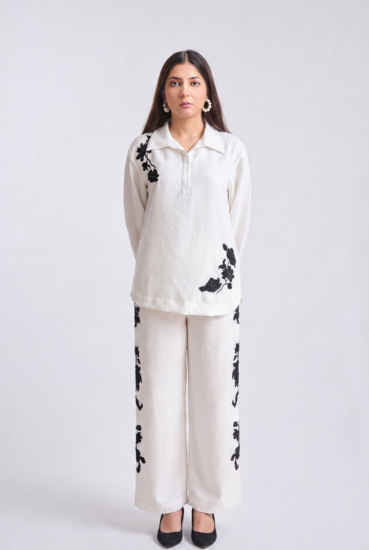 A woman modeling a matching two-piece outfit consisting of a long-sleeve collared top and wide-leg trousers in off-white, both featuring black floral embroidery, set against a plain light gray studio background.