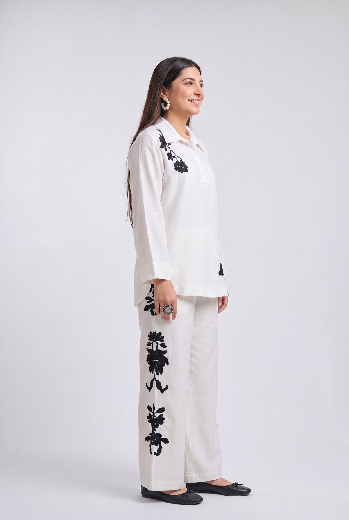 A woman modeling a white two-piece kurta set with black floral embroidery, standing in profile against a light gray studio background.